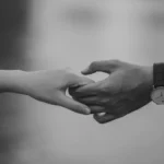 Duas pessoas dando as mãos com delicadeza, em preto e branco, simbolizando conexão emocional e relacionamento íntimo.