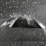 Guarda-chuva preto visto de cima sendo atingido por forte chuva, com gotas respingando ao redor, simbolizando proteção, limpeza emocional e o significado de sonhar com chuva.
