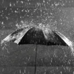 Guarda-chuva preto visto de cima sendo atingido por forte chuva, com gotas respingando ao redor, simbolizando proteção, limpeza emocional e o significado de sonhar com chuva.