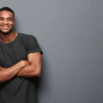 Homem jovem e atraente sorrindo de braços cruzados em frente a uma parede cinza, transmitindo confiança e simpatia.