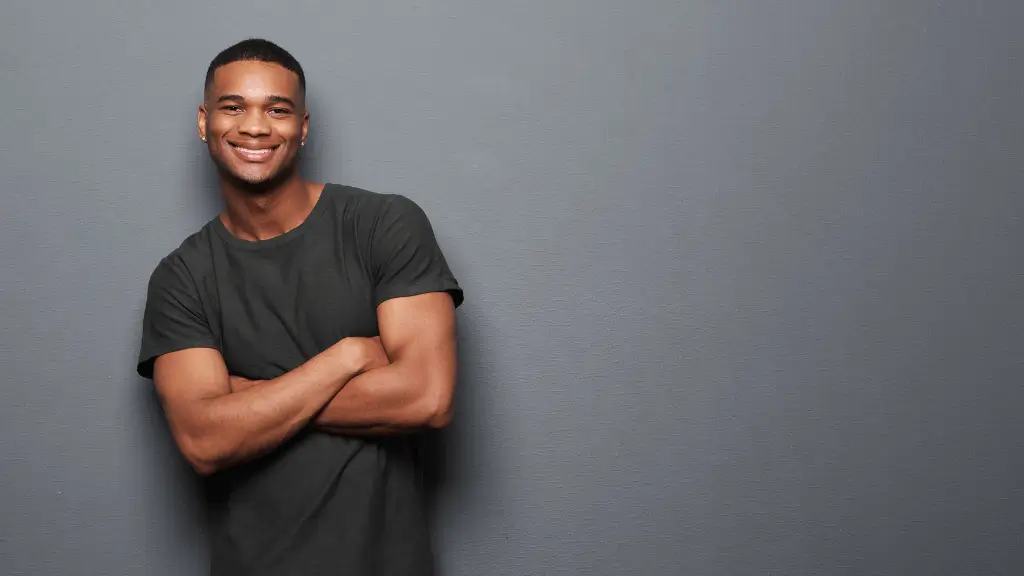 Homem jovem e atraente sorrindo de braços cruzados em frente a uma parede cinza, transmitindo confiança e simpatia.