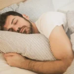 Homem dormindo tranquilamente abraçado ao travesseiro, coberto por lençóis listrados em um quarto aconchegante.