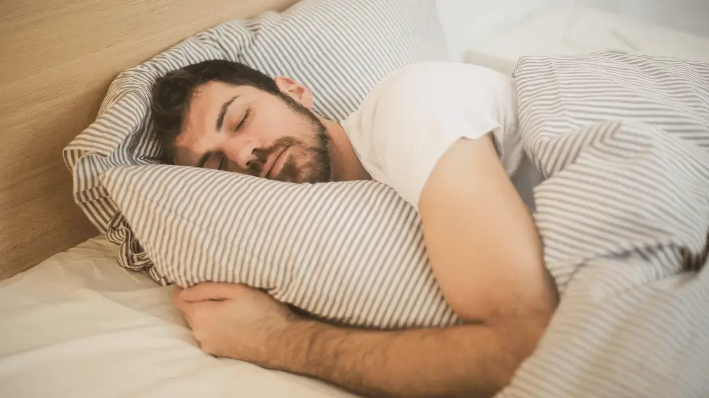 Homem dormindo tranquilamente abraçado ao travesseiro, coberto por lençóis listrados em um quarto aconchegante.